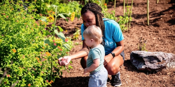 Teacher crouching with a young toddler near sunflowers in an outdoor garden – The Learning Tree Academy in Toccoa, Georgia